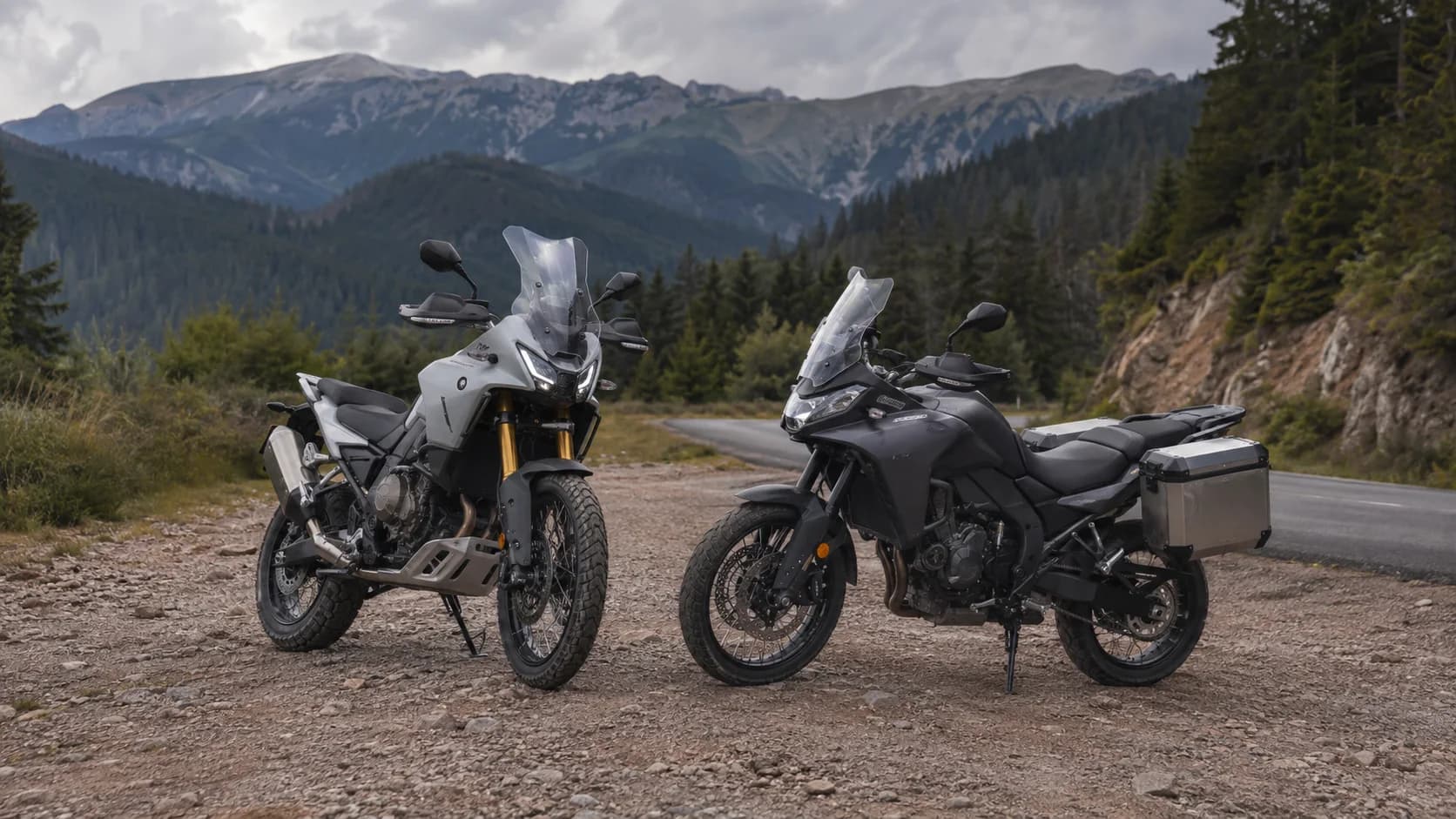 Două motociclete adventure fără sigle oprite pe macadam lângă un drum de munte