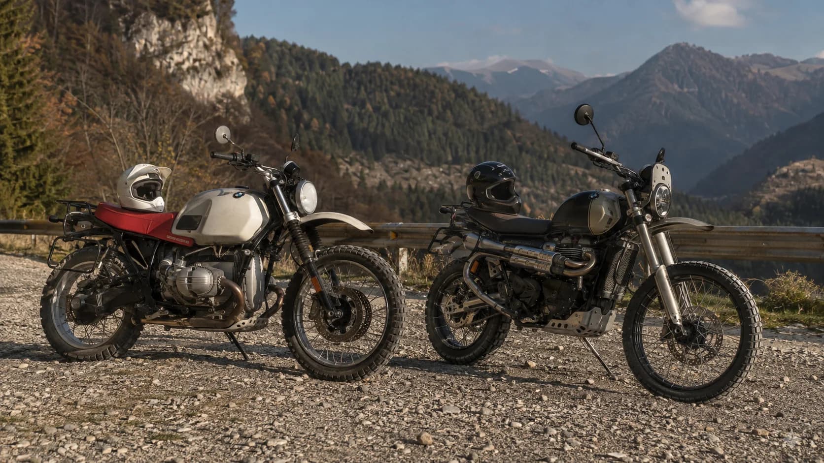 Două motociclete retro adventure parcate pe pietriș lângă un drum de munte